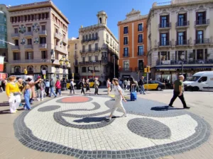 A sunny day in Barcelona, travellers walk across the Joan Miró mosaic on the floor of Las Ramblas